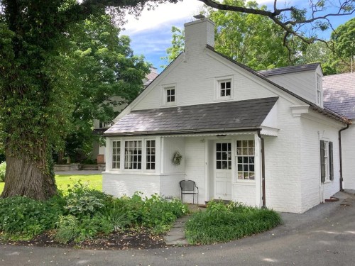 A charming white cottage with a sloped roof, surrounded by greenery and trees on a sunny day.