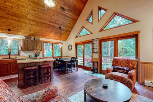 Cozy cabin interior with wooden beams, large windows, and a dining area overlooking a forested view.