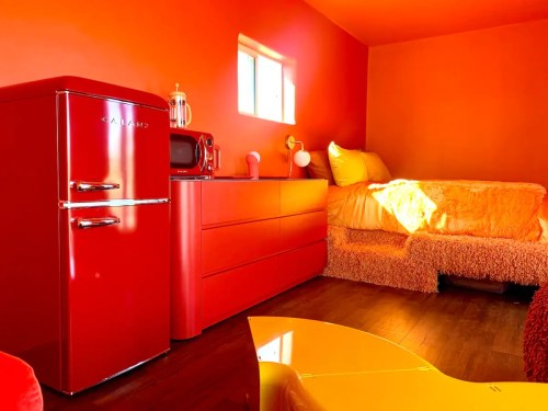 A vibrant room featuring red furniture, including a fridge, dresser, and a cozy bed with orange walls.