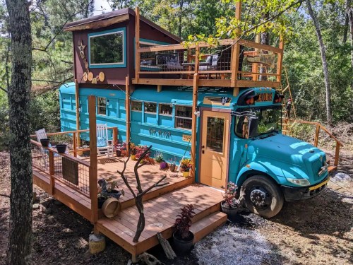 A colorful converted bus with a wooden deck and rooftop terrace, surrounded by trees and potted plants.