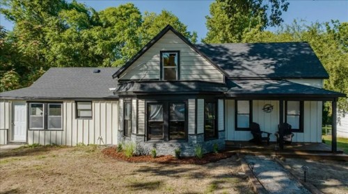 A charming two-story house with a front porch, large windows, and a landscaped yard surrounded by trees.