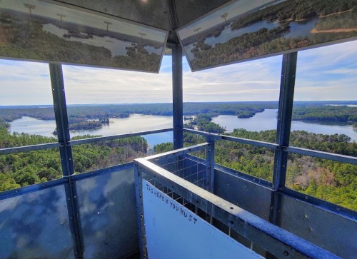 View from a lookout tower overlooking a lake surrounded by lush greenery and distant hills under a clear blue sky.