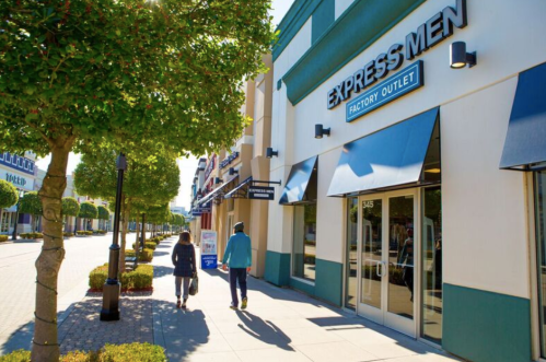 Two people walk along a sunny sidewalk past the Express Men Factory Outlet, lined with trees and shops.