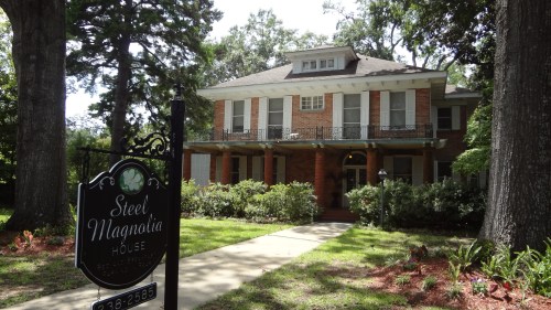 A large, historic brick house with a porch, surrounded by trees and a sign reading "Steel Magnolia House."