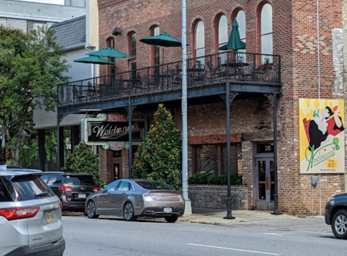 A brick building with a balcony, green umbrellas, and a colorful mural, viewed from the street with parked cars.