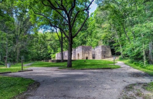 Ruins of a stone structure surrounded by lush greenery and trees, with a pathway leading through the area.