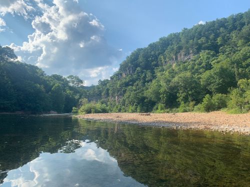 A serene river scene with lush green hills, rocky shore, and fluffy clouds reflecting on the water's surface.