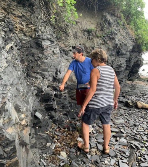 Two people explore a rocky shoreline, examining the geological formations and surrounding greenery.
