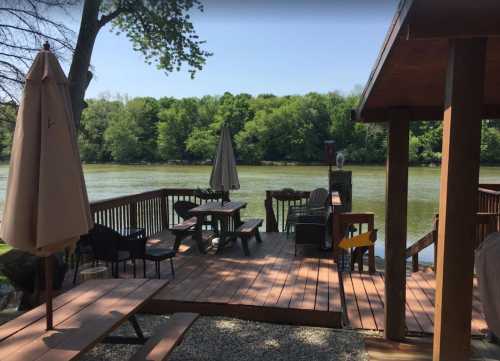 A wooden deck by a river, featuring tables, chairs, and umbrellas, surrounded by lush green trees.