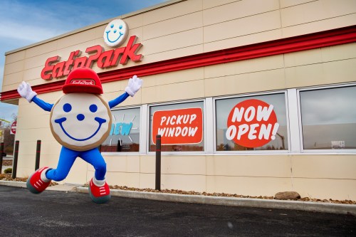 A cheerful mascot in a blue outfit and red cap jumps in front of the EatPark restaurant, showcasing a "Now Open" sign.