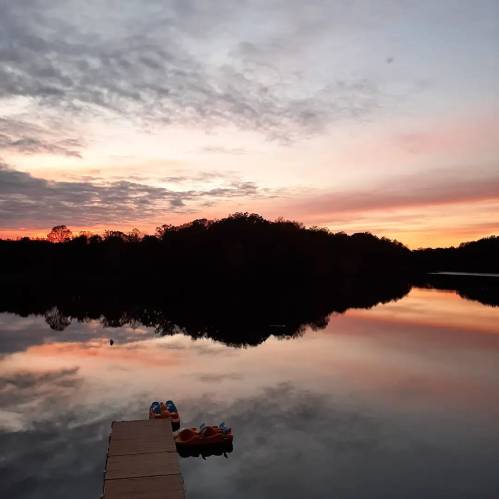 A serene lake at sunset, with a wooden dock and colorful kayaks reflecting on the calm water.