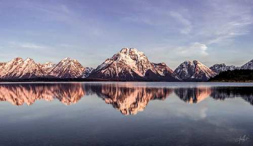 Snow-capped mountains reflect in a calm lake under a clear sky, creating a serene and picturesque landscape.