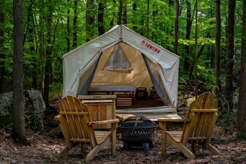 A cozy tent in a forest, with two wooden chairs and a fire pit in front, surrounded by lush greenery.