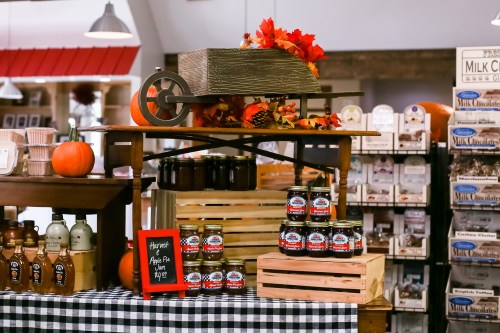 A rustic display featuring a wooden cart with autumn leaves, pumpkins, and jars of apple pie filling on a checkered tablecloth.