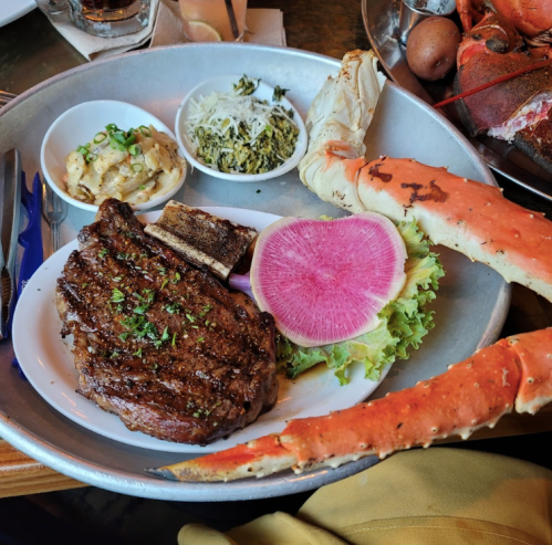 A plate featuring a grilled steak, crab legs, pickled radish, and sides of coleslaw and greens on a metal tray.