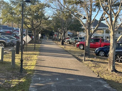 A paved pathway lined with trees, leading past parked cars on one side and buildings in the background under clear skies.