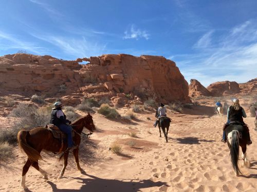 A group of riders on horseback traverses a sandy trail surrounded by red rock formations under a blue sky.