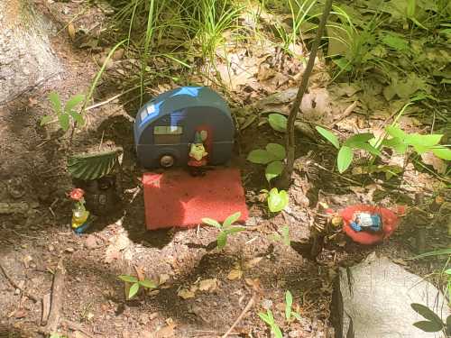 A miniature scene in the woods featuring a blue camper, small figures, and a red mat surrounded by greenery.