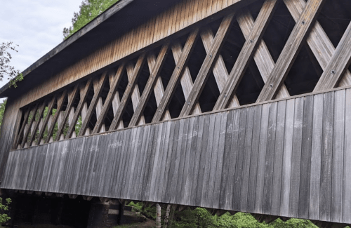 A wooden covered bridge with a diamond-patterned design, surrounded by greenery.