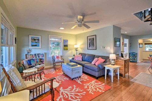 Cozy living room with a blue sofa, coral rug, bookshelves, and natural light from large windows.