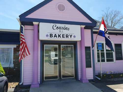 A pink bakery building with columns, an American flag, and a sign reading "Cousins R.I. Bakery."