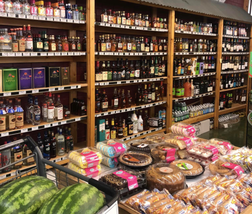 A grocery store aisle filled with various alcoholic beverages on shelves, with baked goods and watermelons in the foreground.