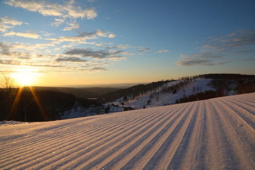 A serene winter landscape with freshly groomed snow, rolling hills, and a sunset casting warm light across the scene.