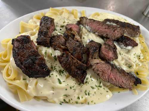 A plate of fettuccine pasta topped with grilled steak pieces and creamy sauce, garnished with herbs.