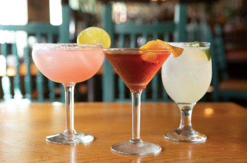 Three colorful cocktails on a wooden table: a pink drink, a martini with orange garnish, and a clear drink with lime.