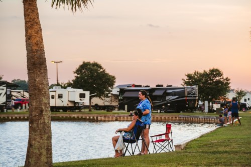 Two people relax by a lake, sitting in chairs with RVs in the background and a sunset sky.