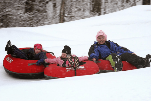 Three people in red snow tubes slide down a snowy hill, smiling and enjoying a winter day.
