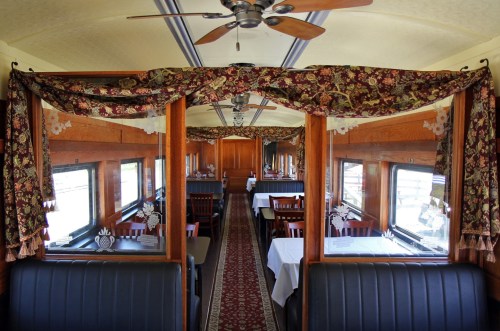 Interior of a vintage dining car with wooden paneling, tables set for dining, and decorative curtains.