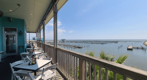 A waterfront restaurant patio with white tables and chairs overlooking a calm bay and docked boats under a clear blue sky.