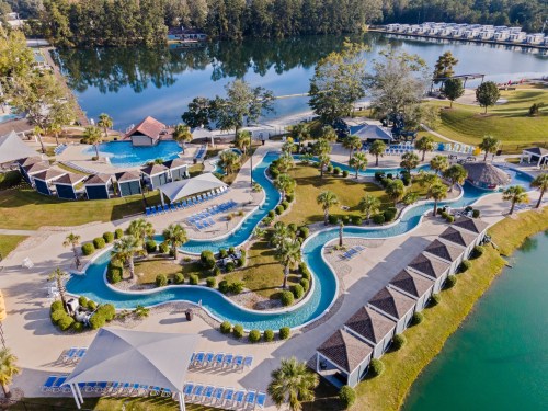 Aerial view of a resort with winding pools, palm trees, and cabins by a serene lake surrounded by greenery.