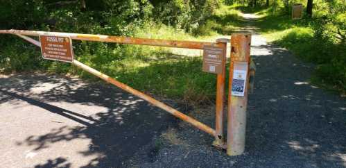 Rusty gate at a trail entrance with signs indicating "Fossil Pit" and information on the area. Lush greenery surrounds the path.