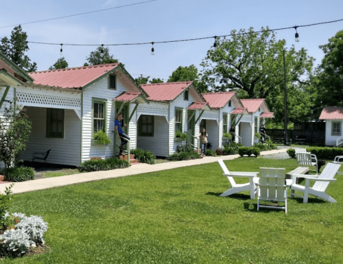 A row of charming white cottages with red roofs, surrounded by green grass and trees, with people enjoying the outdoor space.