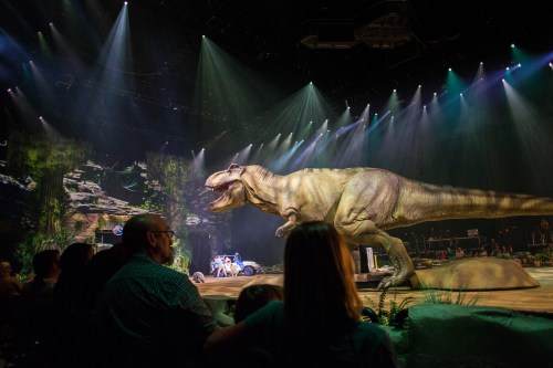 A large dinosaur animatronic performs on stage in a vibrant, illuminated theater, with an audience watching.