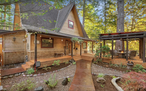 A charming wooden house surrounded by trees, featuring a pathway, garden, and a cozy porch.