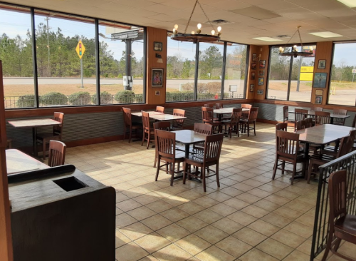 Interior of a restaurant with wooden tables and chairs, large windows, and a view of the outside.