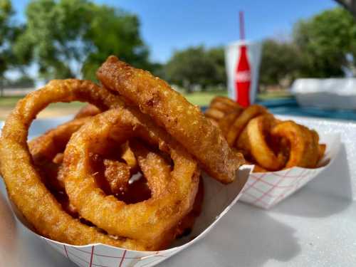 A close-up of crispy onion rings served in a paper tray, with a soft drink in the background and trees in the distance.