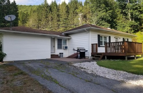 A white single-story house with a wooden deck, grill, and gravel driveway, surrounded by trees.