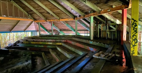 Abandoned indoor space with sloped seating, colorful graffiti, and a wooden roof, illuminated by natural light.
