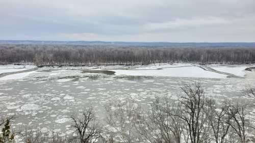 A winter landscape featuring a partially frozen river surrounded by snow-covered trees and a cloudy sky.