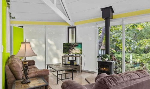 Cozy living space with a TV, lamp, and wood stove, surrounded by large windows and greenery.
