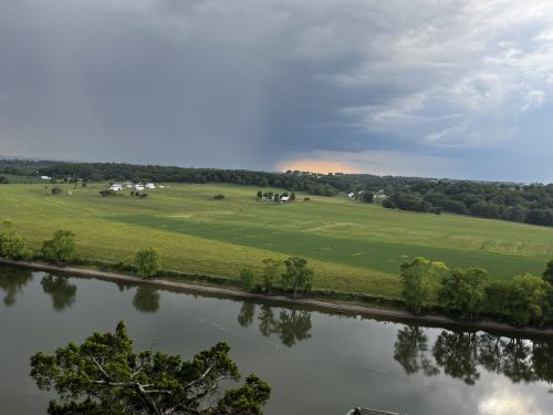 A scenic view of a green field and river under a cloudy sky, with distant farms and a storm approaching.