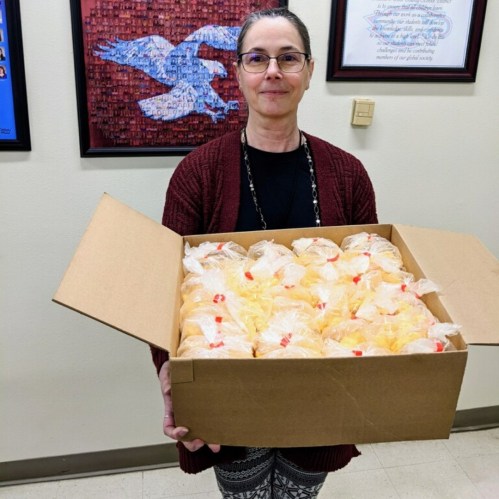 A woman holds a large box filled with individually wrapped items, standing in a room with framed artwork on the walls.