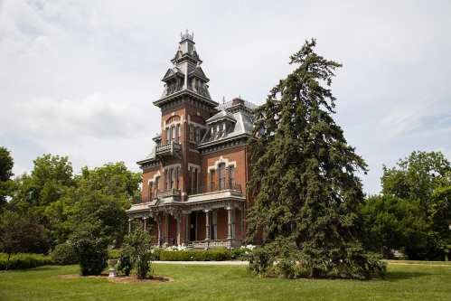 A large, ornate Victorian mansion with a tower, surrounded by lush greenery and trees under a cloudy sky.