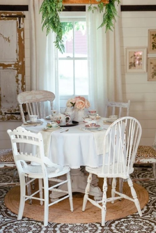 A cozy dining area with a round table set for tea, surrounded by white chairs and decorated with flowers and greenery.