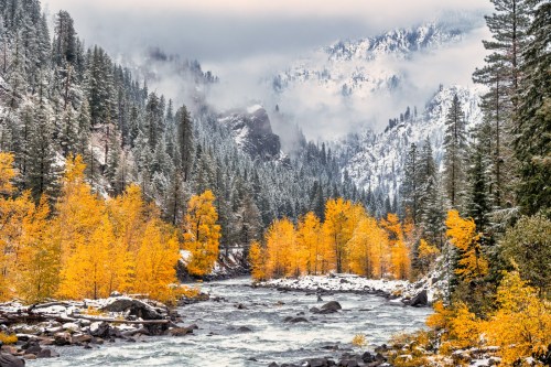A serene river flows through a snowy landscape, surrounded by vibrant autumn trees and misty mountains.