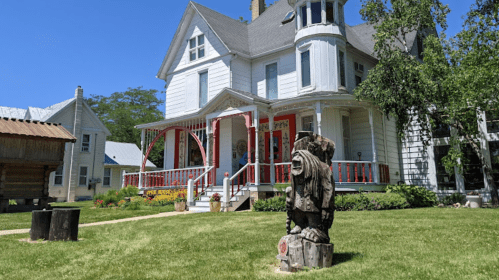 A large white Victorian house with a red porch, surrounded by green grass and a wooden statue in the foreground.